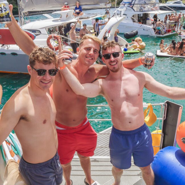Three young men in swim trunks and sunglasses smile and raise drinks while standing on a boat, surrounded by people partying on boats and inflatables in turquoise water on a sunny day.