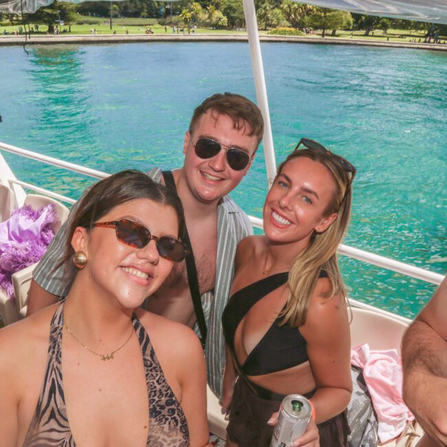 Three people in swimsuits smile at the camera on a boat, with turquoise water and greenery in the background. It’s a sunny day, and everyone looks happy, enjoying a boat party.