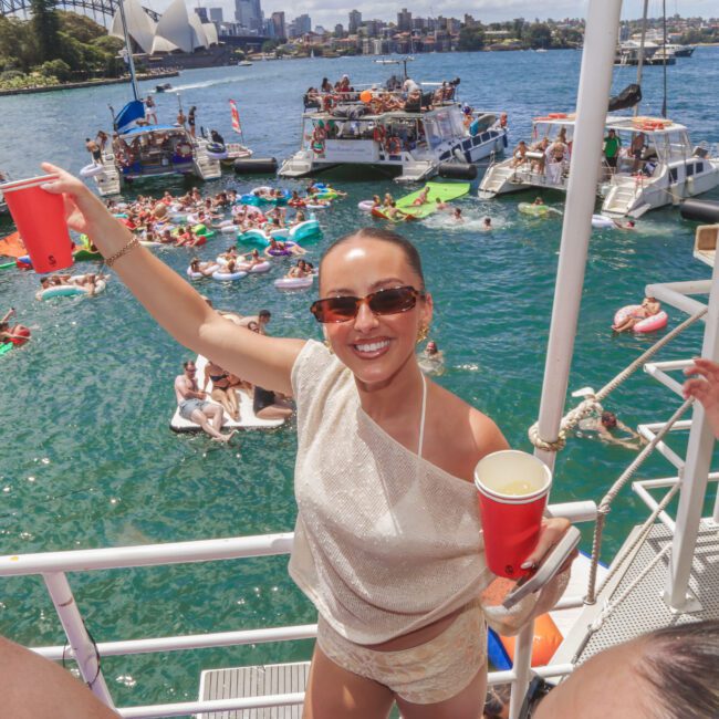 A woman in sunglasses smiles and holds a red cup on a boat, with people swimming, lounging on inflatables, and gathering on nearby boats in a sunny harbor. The Sydney Opera House and city skyline are visible in the background.