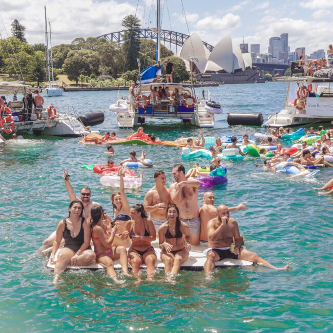 A group of people in swimwear enjoy a floating party on Sydney Harbour, surrounded by boats and others on inflatables, with the Sydney Opera House and city skyline in the background.