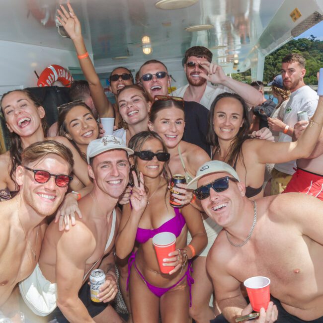 A group of young adults in swimwear pose and smile for a photo on a boat, holding drinks and enjoying a sunny day by clear blue water and lush greenery.