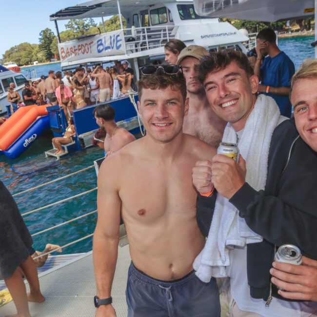 Three smiling young men pose shirtless on a boat, holding drinks, with a lively crowd, inflatable floats, and several boats in the background at a sunny water party.