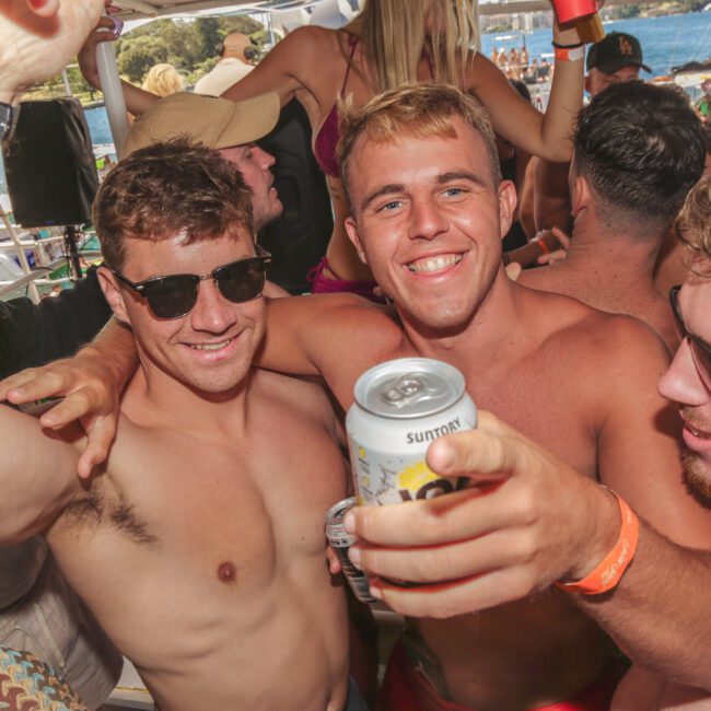 A group of young men in swimsuits smile and pose together at a crowded outdoor party, one holding a can and pointing towards the camera. Sunlight and water are visible in the background.