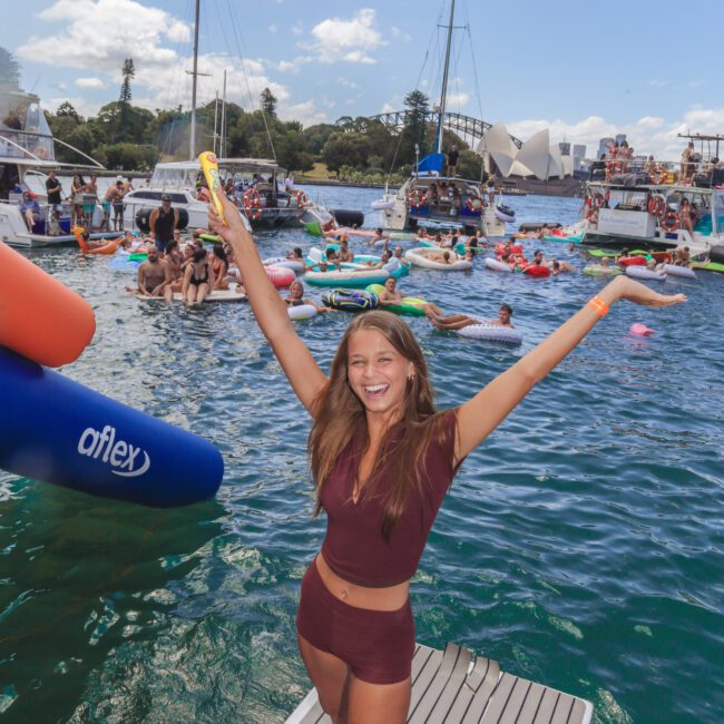 A smiling woman in a burgundy outfit stands on a dock with arms raised, surrounded by boats and people on colorful inflatables, enjoying a sunny day on the water. The atmosphere is festive and lively.