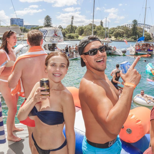 A group of young adults in swimsuits enjoy a sunny day on a boat, smiling and holding drinks, with city buildings, trees, and inflatables visible in the water behind them.