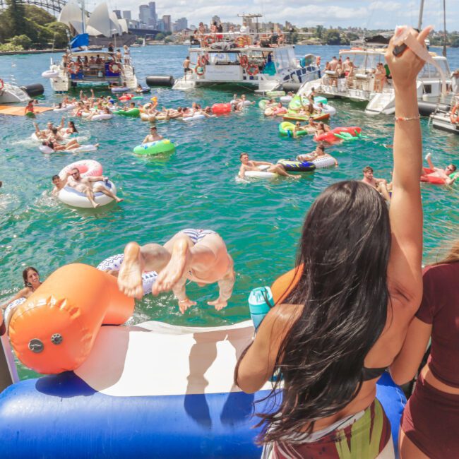 People enjoy a lively boat party on the water, with many swimming, relaxing on floaties, and dancing. Boats are anchored nearby, and one person is mid-air, flipping into the water. Bright, sunny day with a city backdrop.