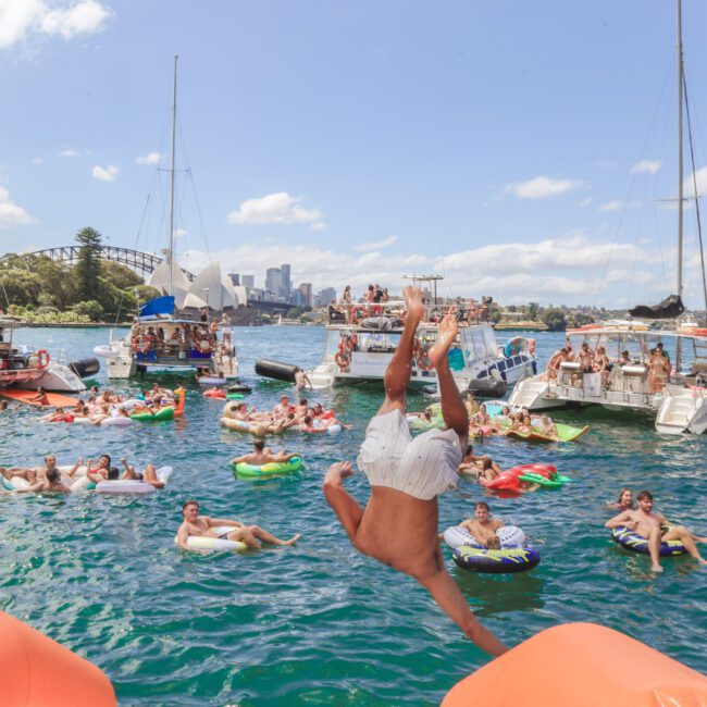 A man flips mid-air above the water while people on inflatables and boats enjoy a lively party on a sunny day in a harbor, with the Sydney city skyline visible in the background.