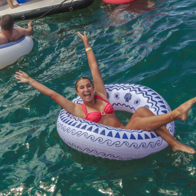 A woman in a pink bikini smiles with arms raised while floating in a blue and white inflatable ring on clear water. Other people relax on inflatables nearby. The scene appears fun and joyful.