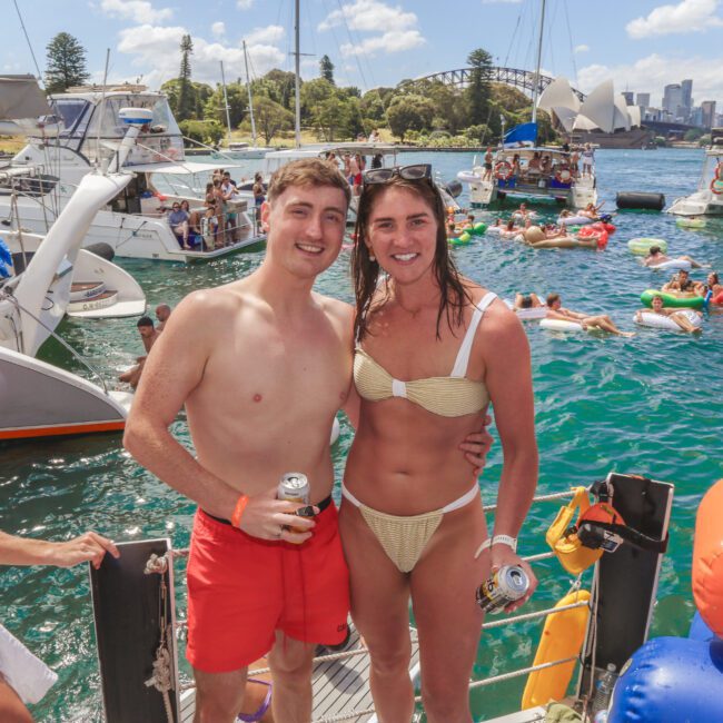 Two people in swimwear smile and pose on a boat dock with drinks in hand. Behind them, many people relax on boats and inflatables in the water on a sunny day, with a city skyline and bridge in the background.