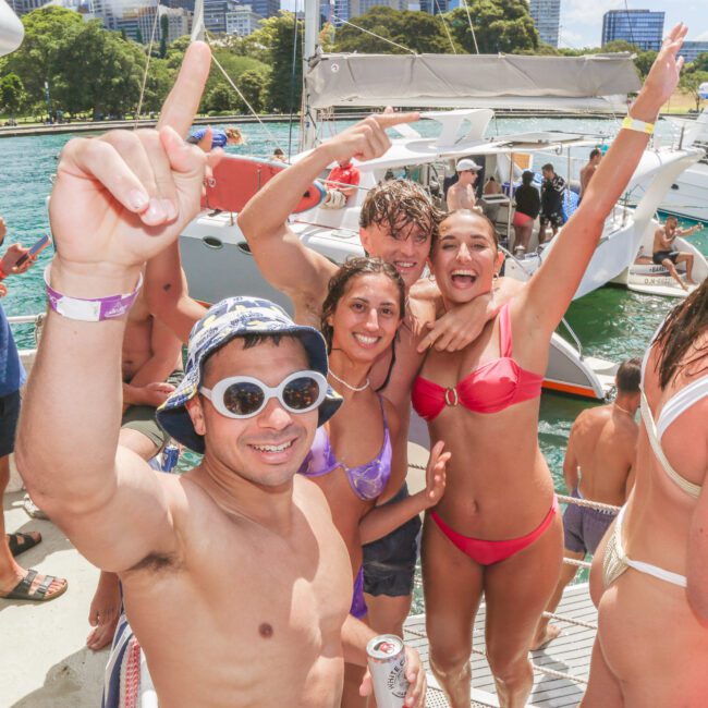 A group of young adults in swimwear smile and celebrate on a boat, with one person pointing upward. Other people and boats are visible on the water in the background. The atmosphere is lively and festive.