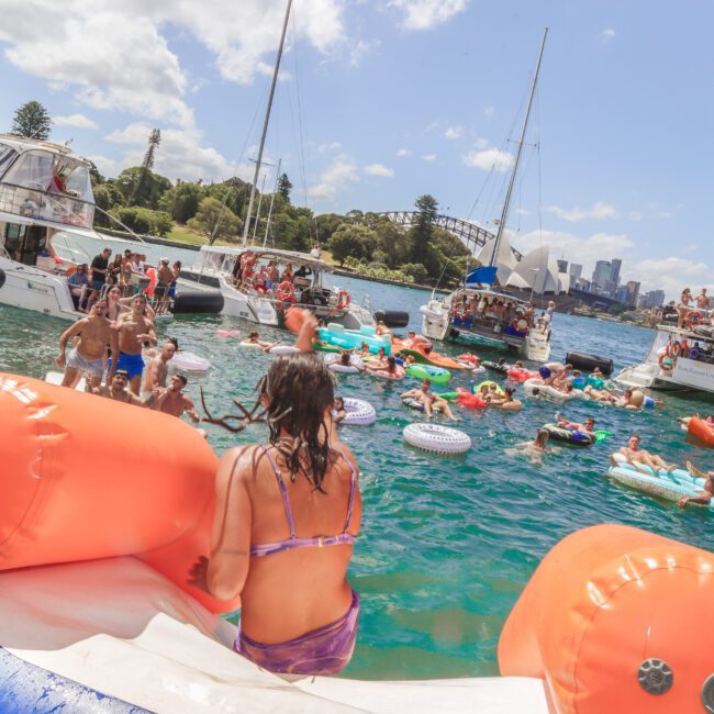 A lively scene on the water shows people in swimsuits swimming, relaxing on colorful inflatables, and socializing near boats. The Sydney Harbour Bridge and city skyline are visible in the background under a partly cloudy sky.