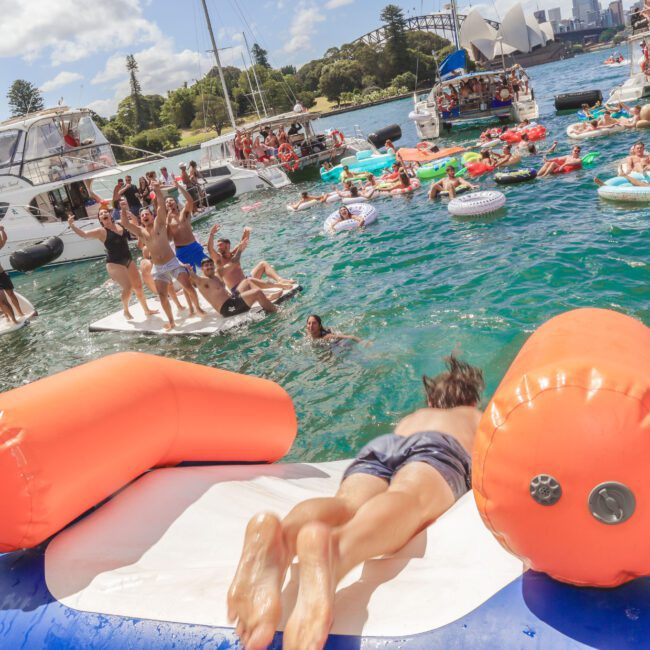 People enjoy a lively party on boats and inflatable floats in a harbor; one person is sliding from an orange and white inflatable into the water. The Sydney Opera House and Harbour Bridge are visible in the background.