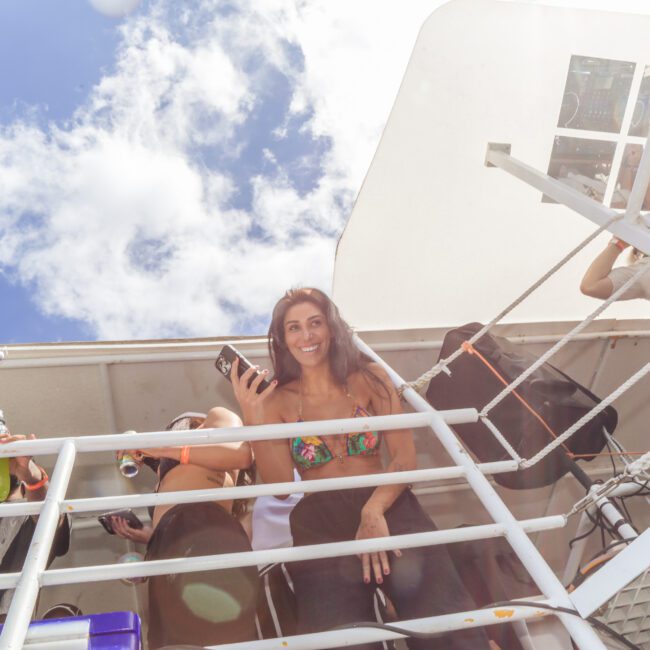 A woman in a bikini top, smiling and holding a phone, stands on the deck of a boat under a partly cloudy sky. Other people relax nearby, some partially visible, with drinks and bags around.