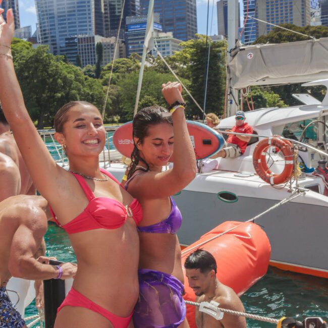Two women in colorful swimsuits dance and smile on a boat with others around them. City skyscrapers and green trees are visible in the background, and the weather is sunny.