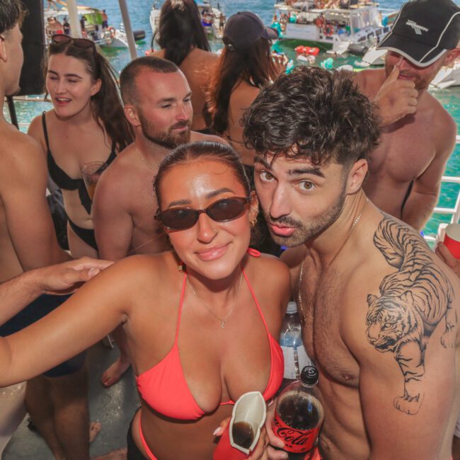 A group of young adults in swimsuits are partying on a yacht, with drinks in hand. The man in front has a tiger tattoo on his shoulder and stands close to a woman in a red bikini. Boats and turquoise water are visible in the background.