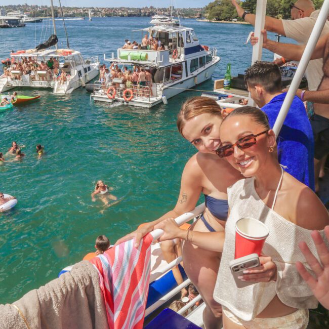 Two women smiling at the camera on a boat, one waving and holding a red cup, with other people swimming and relaxing on floats in the water nearby, and several boats docked in the background.