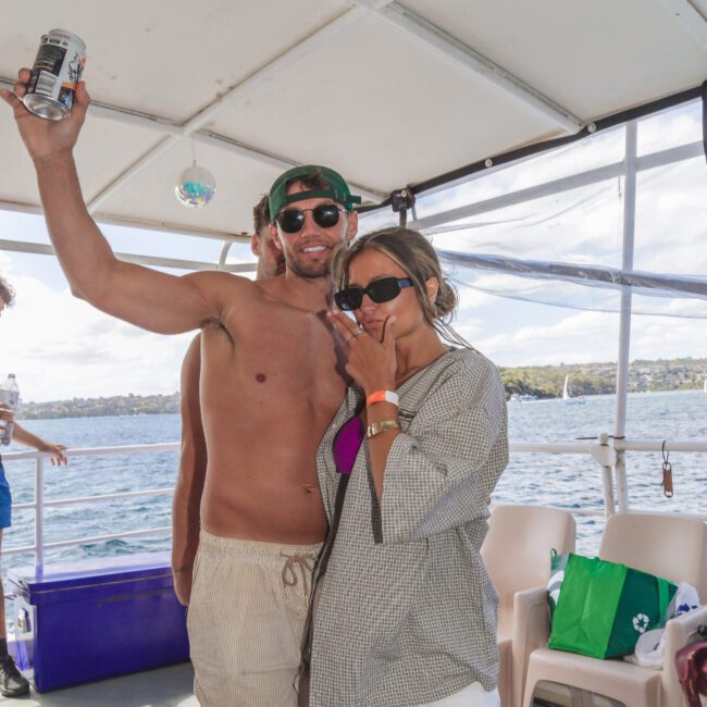 A man in swim trunks and sunglasses holds up a drink, smiling with his arm around a woman in casual clothes and sunglasses on a boat. Seats and water are visible in the background on a sunny day.
