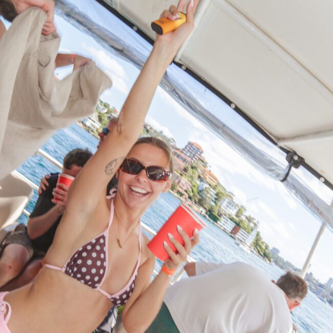 A woman in sunglasses and a polka-dot bikini top smiles and raises her arm while holding a red cup on a boat, surrounded by people enjoying a sunny day on the water.