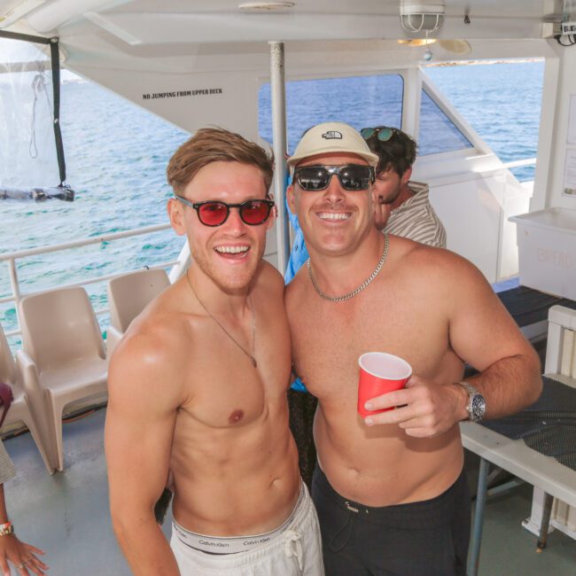 Two smiling men wearing sunglasses and swim trunks pose together on a boat, one holding a red cup. Other people are visible in the background, and the ocean can be seen through the windows.