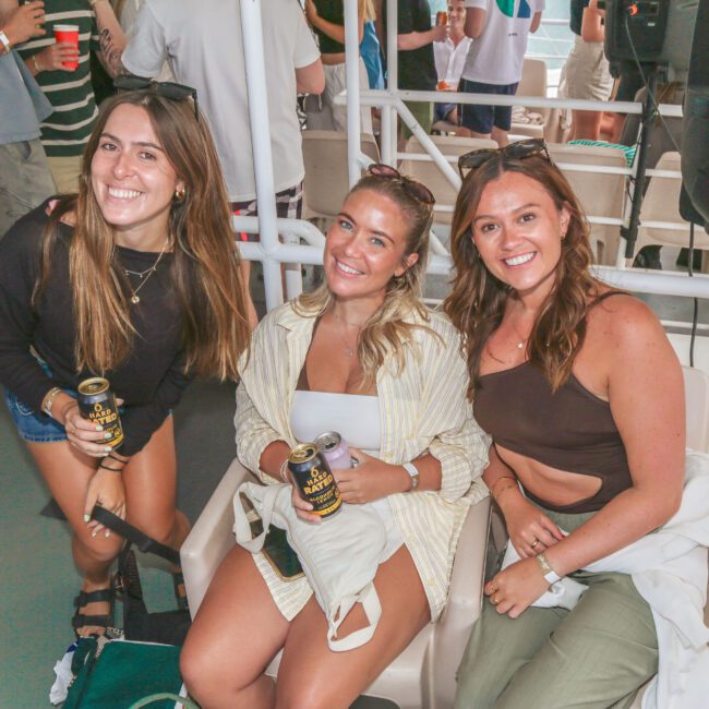 Three young women smiling and sitting together on a boat, holding drinks. People stand and mingle in the background. The atmosphere is casual and lively.
