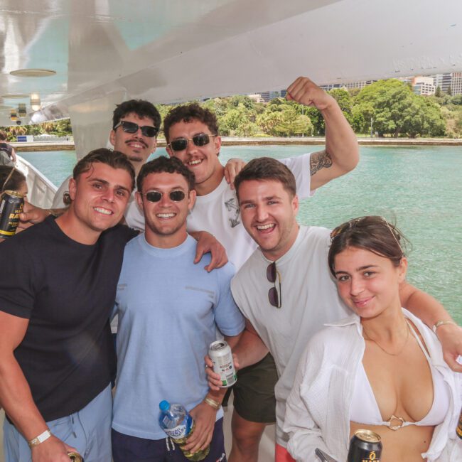A group of six young adults smiling and posing together on a boat, holding drinks. The water and city buildings are visible in the background on a sunny day.