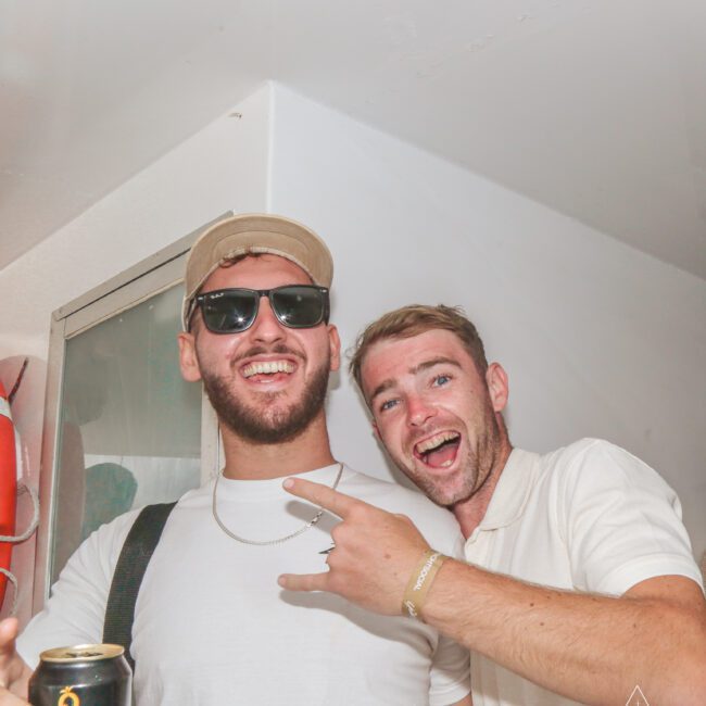 Two men smiling and posing for the camera at a party. One wears sunglasses and a beige cap, holding a can, while the other points at him. Both appear to be enjoying themselves in a brightly lit indoor space.