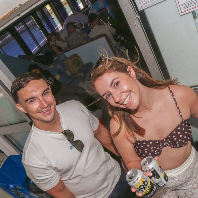 A smiling young man and woman stand close together indoors. The woman holds two cans of drink and wears a polka dot top, while the man wears a white shirt and sunglasses. Other people are seated in the background.