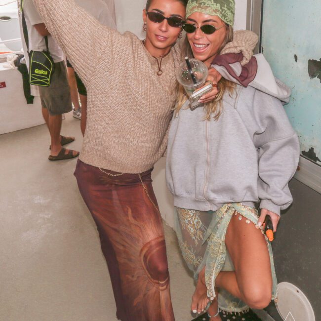 Two women posing and smiling, wearing sunglasses and casual beachwear. One has an arm around the other and holds a round object. There are other people in the background and a bright, relaxed atmosphere.