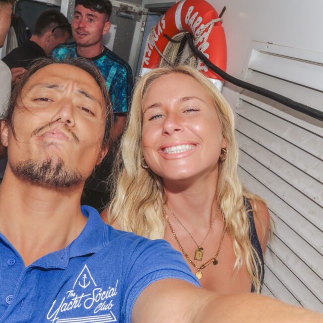A man in a blue "Yacht Social Club" polo shirt takes a selfie with a smiling blonde woman; several people are in the background on what appears to be a boat.