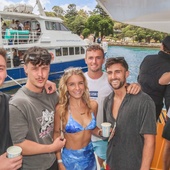 A group of five young adults, four men and one woman, smile together on a boat. Another man drinks from a can nearby. Another boat and water are visible in the background, along with trees and people.