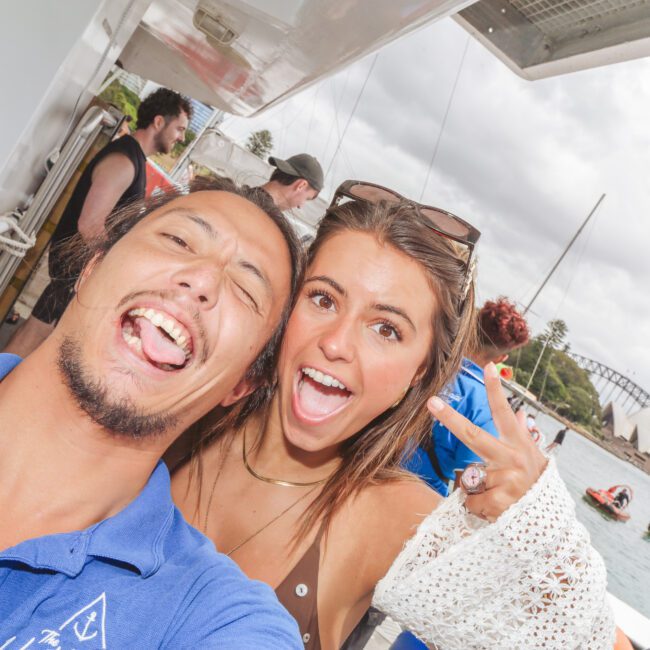 Two people smile and pose for a selfie on a boat, with the Sydney Opera House and Harbour Bridge visible in the background. The woman flashes a peace sign; other people and small boats are in the water behind them.
