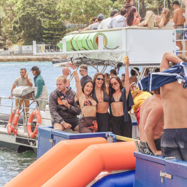 A group of people in swimwear enjoy a sunny day on a boat with an inflatable slide, smiling and posing for photos. Others socialize nearby, and the water and trees are visible in the background.