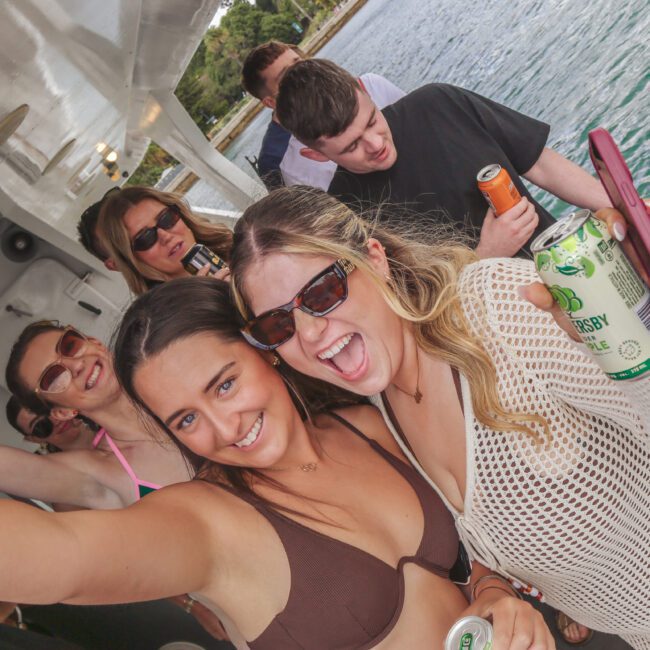 A group of young adults smiling and posing for a selfie on a boat, holding drinks. The background shows water and greenery, and everyone appears to be enjoying a sunny day together.