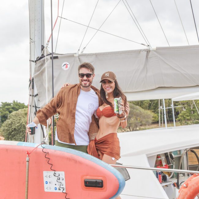 A man and woman smiling and posing on a boat, standing next to a red paddleboard. Both are wearing brown outfits and sunglasses. The woman holds a can and wears a cap. Other people and greenery are visible in the background.