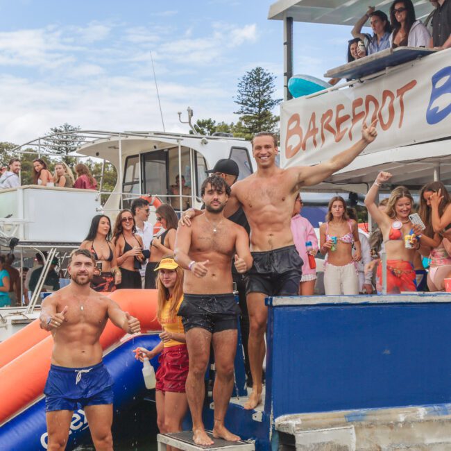 A group of people at a lively boat party with three men and a woman posing on a platform near an inflatable slide. Others are mingling on the boats, with "Barefoot Blue" visible on a sign.