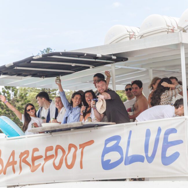 A group of people smile and wave while standing on the deck of a boat named "Barefoot Blue." Some hold drinks, and the atmosphere appears festive and relaxed. The boat is docked near greenery under a partly cloudy sky.
