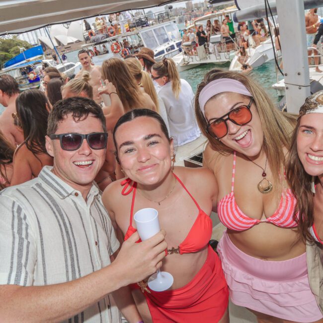 A group of young adults in swimsuits and summer attire smile at the camera during a lively boat party. Other guests mingle in the background near the water, with yachts and greenery visible.