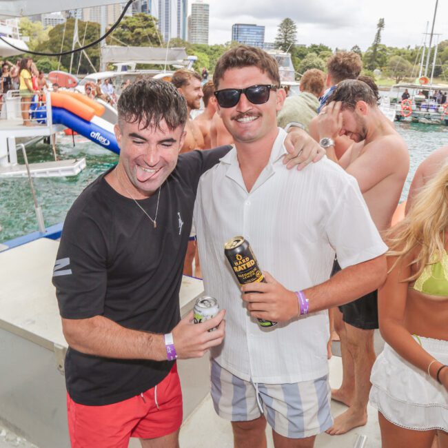 Two men smile and pose with drinks on a boat during a lively party; one has his tongue out. Other partygoers in swimsuits socialize in the background near water and green trees.