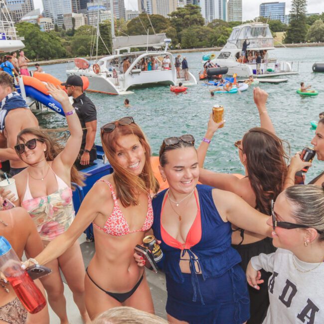 A group of young adults in swimwear enjoy a lively boat party, laughing, dancing, and holding drinks. Other boats and people on floaties are visible in the water, with a city skyline and trees in the background.