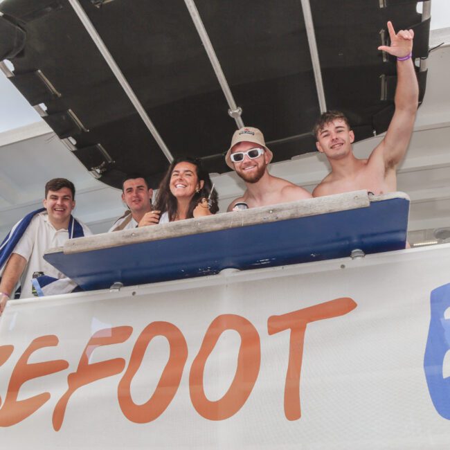 Five smiling people, some shirtless, stand on a boat deck behind a banner that reads "BAREFOOT." One person raises a hand in a celebratory gesture. The sky is partly cloudy in the background.
