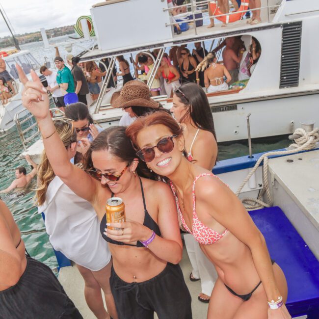 A group of young women in swimsuits smile and pose for a photo on a crowded party boat, with other people and boats in the background on the water. One woman holds a drink and raises her hand in celebration.