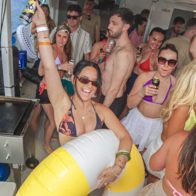 A group of people in swimsuits enjoy a lively boat party. A woman in sunglasses and a bikini smiles and raises her arm, holding a drink and a yellow-and-white pool float. Others are chatting, dancing, and holding drinks.