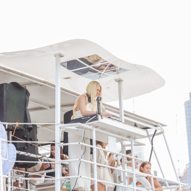 A woman with blonde hair stands behind a DJ booth on the upper deck of a white boat, performing for a group of people. Modern city buildings are visible in the background.