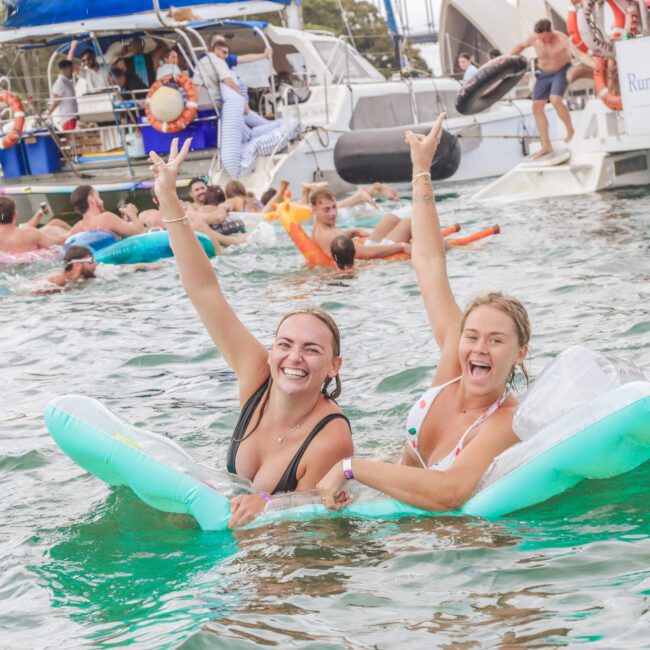 Two women in swimsuits smile and throw up peace signs while lounging on inflatable floats in the water near party boats with people celebrating in the background. The scene is lively and joyful.