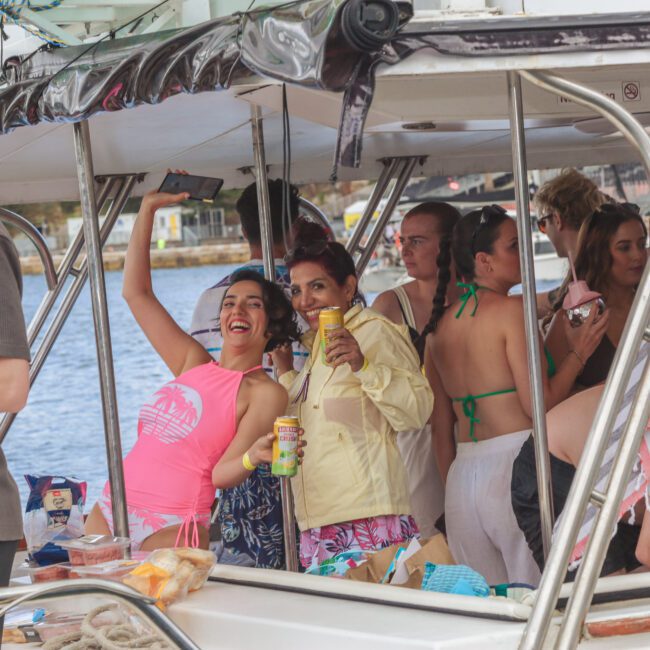 A group of people in swimwear enjoy drinks and snacks on a boat. One woman in a pink top takes a selfie, while others smile and chat. The boat is docked near the water, and snacks are spread out on the counter.
