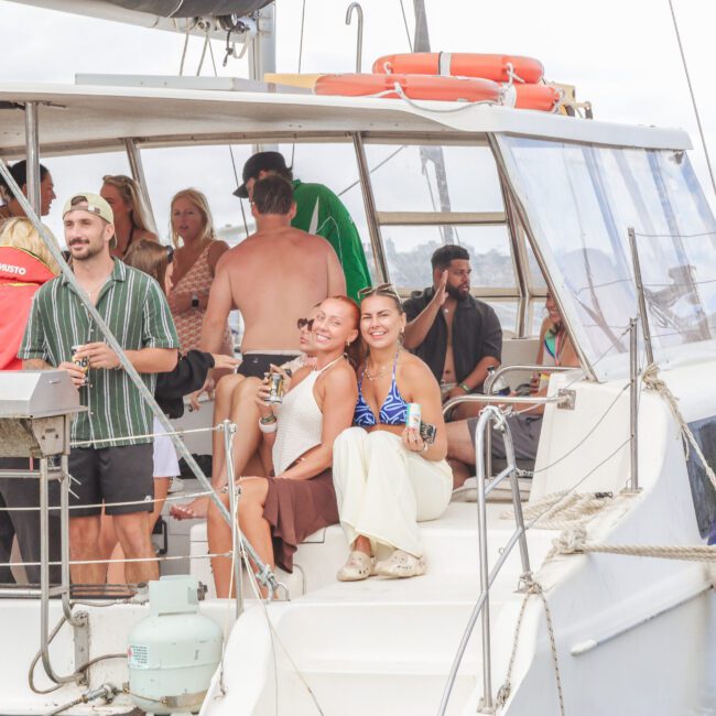 A group of people enjoy a party on a white boat. Two women sit smiling at the front, holding drinks, while others stand and chat in the background. The boat is docked near the shore on a partly cloudy day.