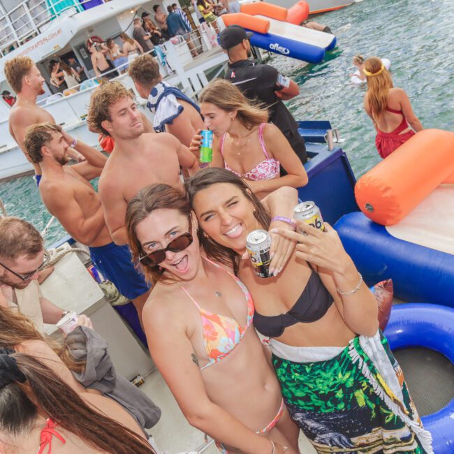 A group of young adults in swimsuits enjoy a lively boat party, holding drinks and smiling at the camera. Others mingle nearby and swim in the water, with boats and inflatables in the background.