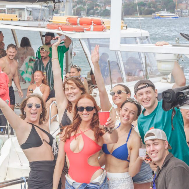 A group of young adults in swimsuits and summer clothing smile and pose for the camera on a boat, holding drinks. The background shows water, other boats, and a distant shoreline with buildings.