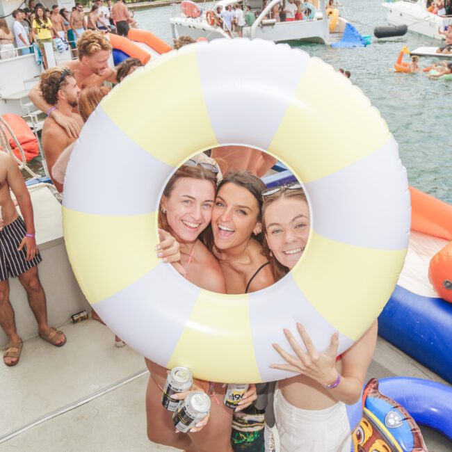 Three smiling women pose through a yellow and white inflatable ring on a boat at a lively pool party, surrounded by friends, floats, and water in the background.