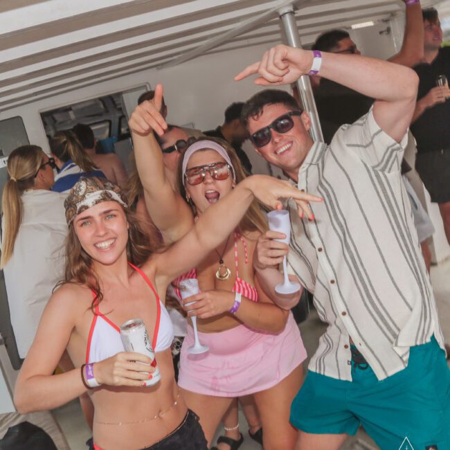 A group of people in swimwear smile, pose, and celebrate on a boat, holding drinks and dancing. The atmosphere is lively and festive, suggesting a summer party or social event.